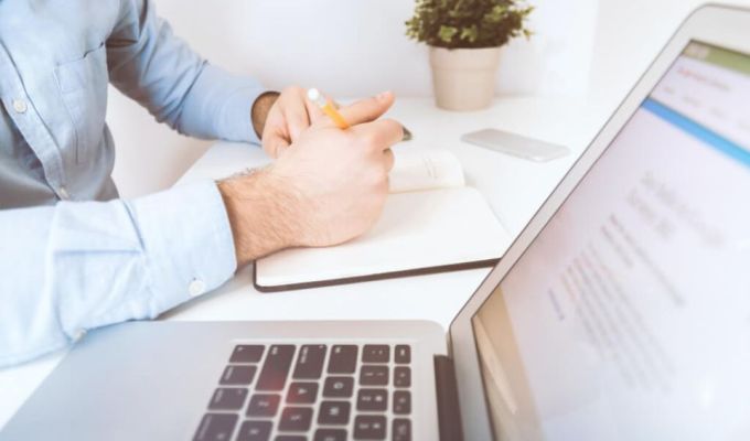 Obrazek dekoracyjny:osoba notuje cos w notatniku, obok jest włączony laptop